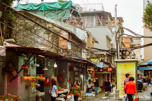 A busy street in Guangzhou lined with older one-, two-, and three-story buildings, stalls selling fruits and vegetables, and shoppers passing by.
