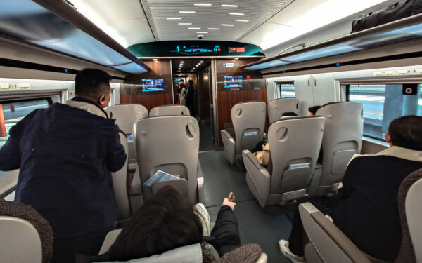Viewed from behind, several passengers are sitting or standing in the cabin of a sleek, high-speed train departing from Beijing. In each row and on each side of the aisle, there are two large seats with gray seat-backs and overhead shelves for storing luggage. The front of the cabin has two digital display screens set into dark wood paneling; the next cabin is visible ahead, through the open door.