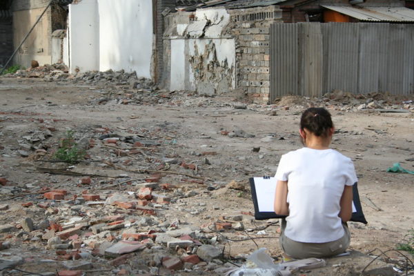 Her back to the camera, a young woman sits amid bricks and rubble with her sketchpad, drawing the site of a recently demolished building.