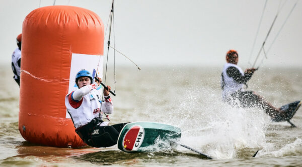 A kiteboarder from the Chinese team—wearing a black wetsuit and a blue helmet—grips a bar attached to the kitefoil above him, leans close to the surface of the water, tips his board, and sends up a spray of water as he maneuvers past a large orange buoy.