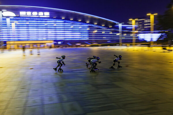 Four young skaters wearing roller skates, helmets, and other safety equipment crouch low as they race across a vast empty concrete square in front of Beijing's National Stadium. It is nighttime, and the blue and white lights of the stadium behind them and the amber lights above them in the square are very blurred.