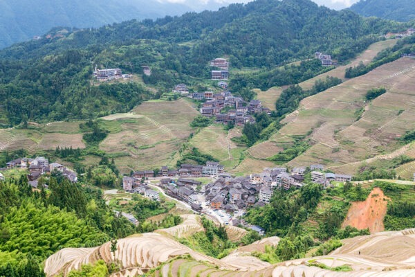 A gorgeous aerial view of a valley in Dazhai, Guangxi province, features clusters of gray, white, and reddish buildings nestling among intricately terraced hillsides and heavily forested hilltops.