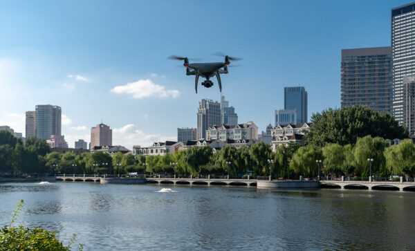 An insect-like drone with two glowing red lights like eyes hovers over a scenic lake, surrounded by tall buildings, in Dalian, China.