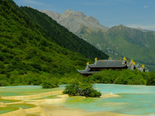 A series of pale turquoise travertine pools lead the eye toward a temple complex with tiled rooftops and yellow roof ornaments, and beyond that, the forested slopes and dramatic towering peaks of Sichuan’s Huanglong National Park.