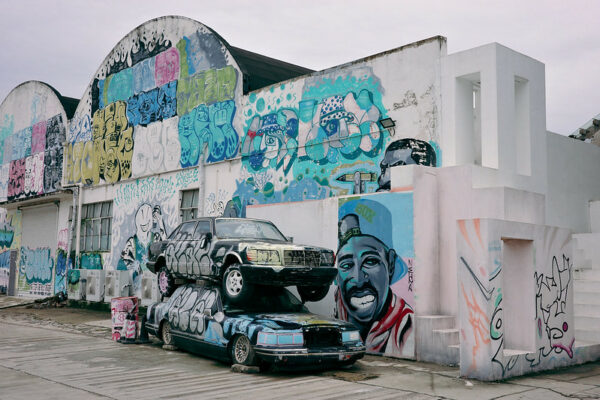 Two beat-up and graffiti-covered cars, one stacked atop the other, have been placed like a sculptural installation in front of an industrial building covered with colorful, artistic graffiti portraits, texts, and symbols.