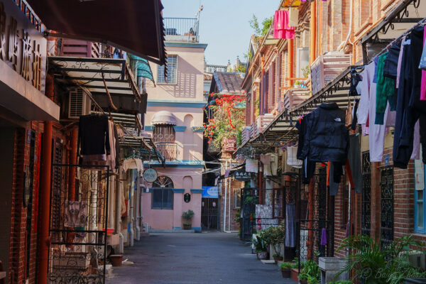 The two-story dwellings on either side of a narrow alleyway in Xiamen are adorned with decorative plants and hung with laundry. At the far end of the alleyway is a three-story building with a cheerful pink and turquoise facade and a rooftop balcony with metal railings.