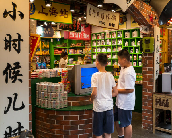 Two boys in white t-shirts and blue shorts play a video game on an old-school television set at the entrance of a small and pleasantly cluttered shop selling various vintage Chinese products from decades past.