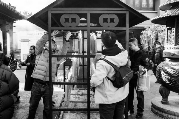 Several visitors to a temple light long sticks of incense from a bank of candles arranged beneath a metal stand with a sloping roof. Behind them in the temple courtyard are other visitors, all dressed in winter jackets, walking around or making offerings of incense.