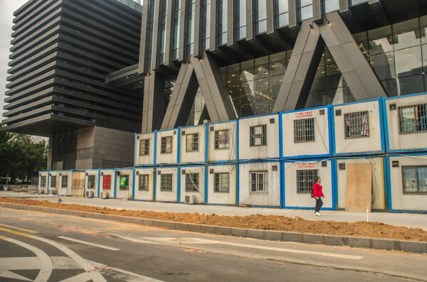 Along the sidewalk, at the foot of a gleaming glass and steel skyscraper, a series of small boxy rooms with barred windows are spartan housing for migrant construction workers.