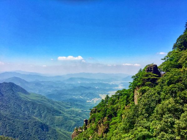 Below a bright blue sky, and beyond the dramatic, deeply forested peaks, a series of verdant hills undulate into the distance, seeming to merge with a bank of clouds on the horizon.